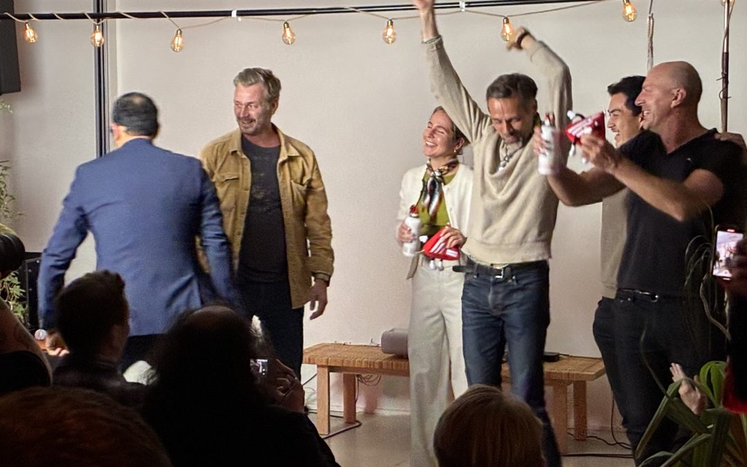 Six people on a small stage celebrating, one with an arm raised in the air, holding prizes, in front of a string of warm lights